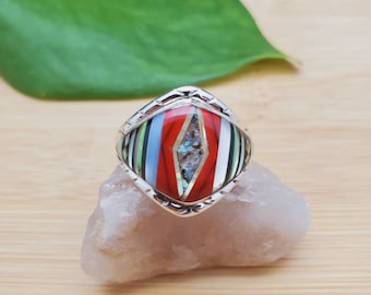 Op de afbeelding: Een zilveren ring met een vierkante inleg van rode, blauwe, groene en witte stenen. De ring ligt op een witte steen.