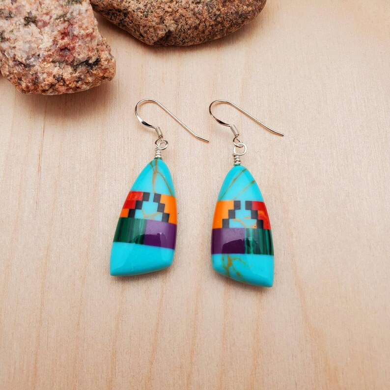 May include: A pair of turquoise dangle earrings with a geometric inlay of red, orange, green, purple, and black stones. The earrings are made of sterling silver.