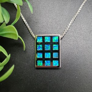 Peut inclure: Collier argenté avec un pendentif carré orné d'une grille de douze pierres d'opale bleu-vert. Le pendentif est serti sur un fond noir, et la chaîne est de style maillon délicat. Le collier est présenté sur un fond sombre.