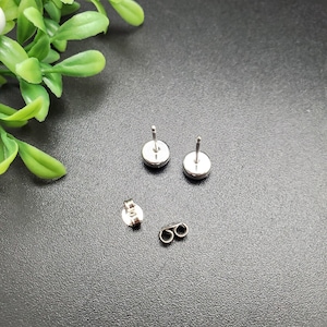 May include: A pair of silver stud earrings with a round, flat design. The earrings are shown on a dark gray surface.