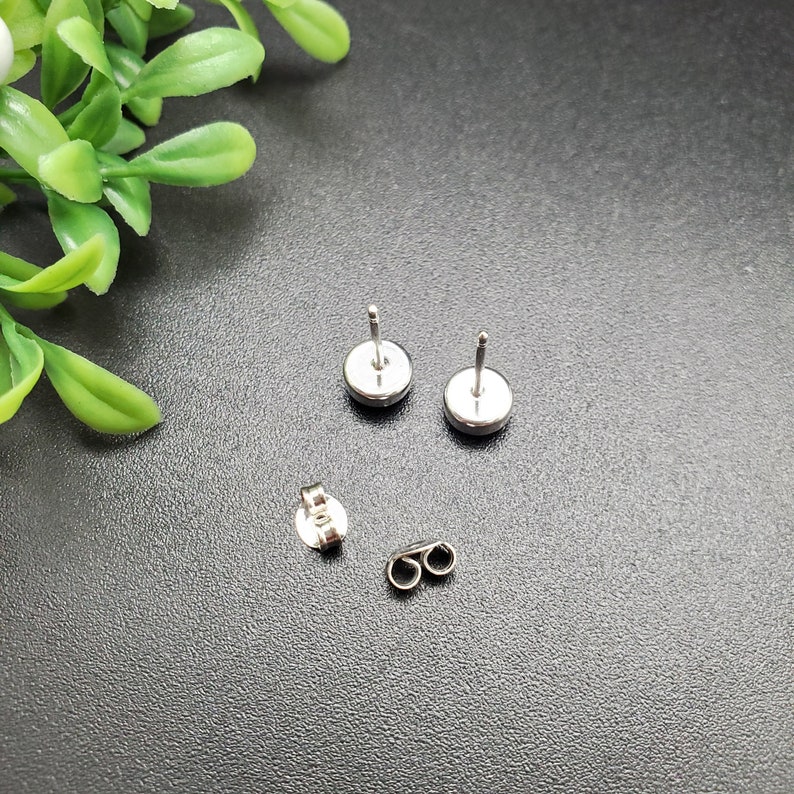 May include: A pair of silver stud earrings with a round, flat design. The earrings are shown on a dark gray surface.