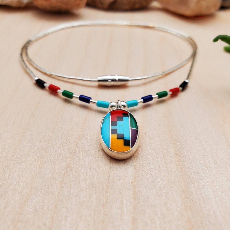 Puede incluir: Un collar de plata con un colgante ovalado multicolor. El colgante est&aacute; hecho de turquesa, rojo, amarillo, verde, negro y morado. El collar tiene un cierre magn&eacute;tico.