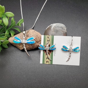 May include: A silver necklace and earrings set featuring a dragonfly pendant and earrings with blue opal accents. The jewelry is displayed on a card with the text "Silver Silver".