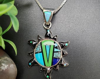 May include: A silver necklace with a sunburst pendant. The pendant features a black border with turquoise, green, and blue inlay. The pendant is approximately 2 inches in diameter and hangs from a silver chain.