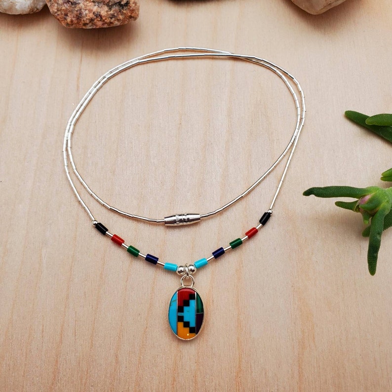 Puede incluir: Un collar de plata con un colgante ovalado multicolor. El colgante est&aacute; hecho de turquesa, rojo, amarillo y piedras negras. El collar tiene un cierre magn&eacute;tico.