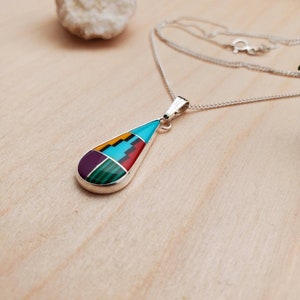 Puede incluir: Un collar de plata con un colgante en forma de l&aacute;grima con una incrustaci&oacute;n multicolor de turquesa, rojo, amarillo, verde y morado.