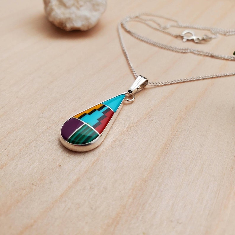 Puede incluir: Un collar de plata con un colgante en forma de l&aacute;grima con una incrustaci&oacute;n multicolor de turquesa, rojo, amarillo, verde y morado.