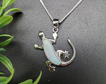 Peut inclure: Un collier avec pendentif en argent représentant un lézard. Le lézard a un centre en forme d'opale et est suspendu à une fine chaîne en argent. Le pendentif est détaillé avec des pattes, une queue et une tête. Le fond est sombre.