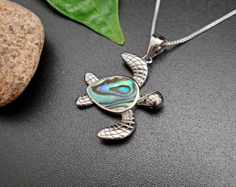 Puede incluir: Un collar con un colgante de tortuga de plata con una incrustación de concha de abulón verde, azul y morado. El collar está sobre un fondo negro.