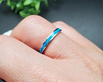 Pode incluir: Um anel de prata com uma faixa de opala azul brilhante. A opala é embutida em um canal fino, criando um contraste vibrante com a prata. O anel é mostrado em um dedo, com um fundo desfocado.