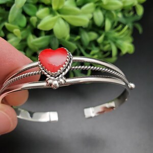 Op de afbeelding: Een zilveren armband met een rood hartvormig steentje in het midden. De armband heeft een gedraaid draadontwerp en is versierd met kleine zilveren kralen.