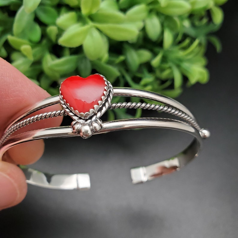 Op de afbeelding: Een zilveren armband met een rood hartvormig steentje in het midden. De armband heeft een gedraaid draadontwerp en is versierd met kleine zilveren kralen.