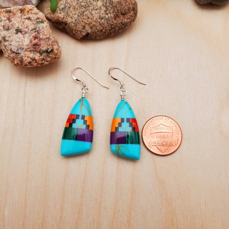 May include: A pair of turquoise dangle earrings with a geometric pattern of green, purple, orange, and black stones. The earrings are approximately the size of a US penny.