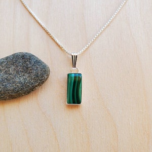 Puede incluir: Un collar de plata con un colgante rectangular de malaquita verde vibrante con rayas verde oscuro. El colgante cuelga de una delicada cadena de plata. El fondo es una superficie de madera clara.
