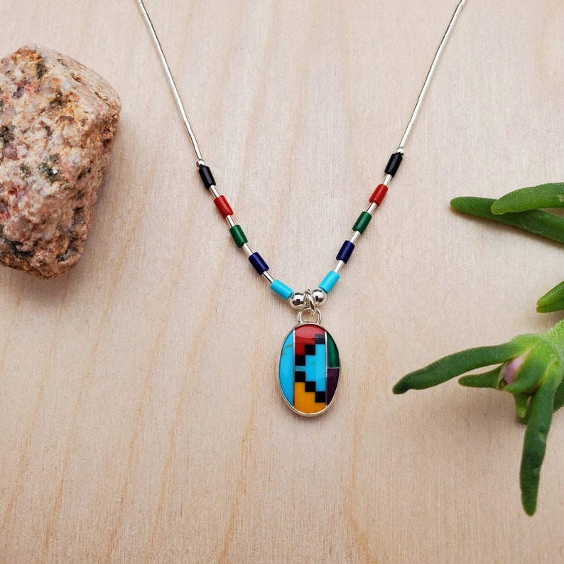 Puede incluir: Un collar de cadena de plata con un colgante ovalado multicolor. El colgante est&aacute; hecho de turquesa, rojo, amarillo, negro y verde. La cadena est&aacute; decorada con peque&ntilde;as cuentas de los mismos colores.