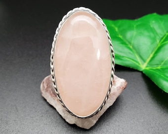 May include: A silver ring with a large oval-shaped pink rose quartz gemstone. The ring has a rope-like design around the edge of the setting.