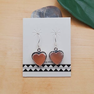 May include: A pair of silver heart-shaped earrings with pink abalone shell inlays. The earrings are displayed on a white card with a black and white geometric pattern.
