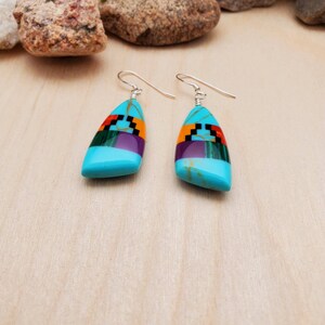 May include: A pair of turquoise dangle earrings with geometric inlay designs in shades of purple, green, red, and black. The earrings are made of sterling silver and feature a teardrop-shaped turquoise stone.