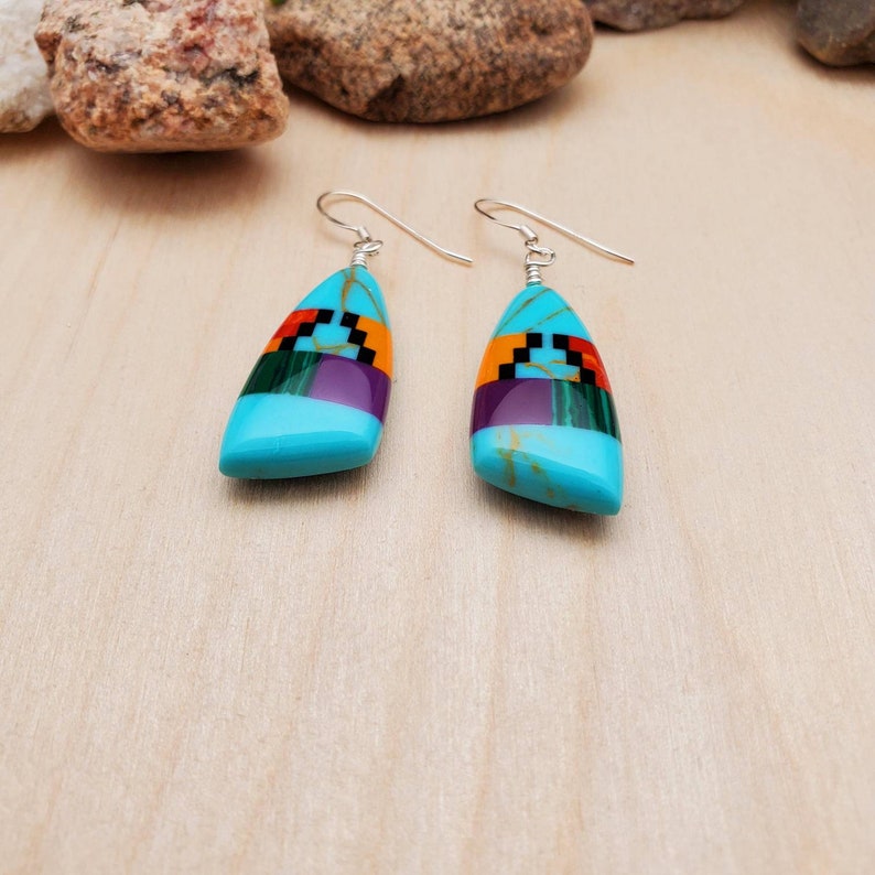 May include: A pair of turquoise dangle earrings with geometric inlay designs in shades of purple, green, red, and black. The earrings are made of sterling silver and feature a teardrop-shaped turquoise stone.