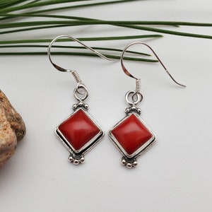 May include: A pair of silver earrings with red square-shaped stones. The earrings have a simple design with a small silver bead detail at the bottom.