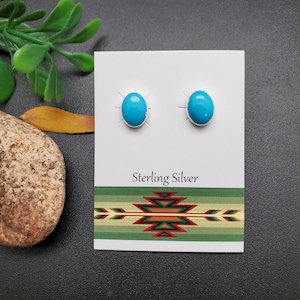 Puede incluir: Un par de pendientes de plata de ley con piedras turquesa. Los pendientes son de forma ovalada y tienen un engaste de plata. Las piedras turquesa son de un color azul brillante.