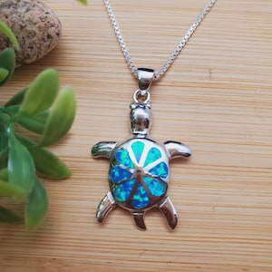 Könnte beinhalten: Eine silberne Schildkröten-Anhänger-Kette mit einem blauen Opal-Inlay. Die Schildkröte ist nach vorne gerichtet, mit erhobenem Kopf und ausgestreckten Beinen. Der Opal ist leuchtend blau mit einem grün schillernden Schimmer.