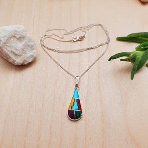 Puede incluir: Un collar de cadena de plata con un colgante en forma de l&aacute;grima. El colgante est&aacute; hecho de plata e incrustado con piedras turquesa, rojas, amarillas, verdes y moradas.