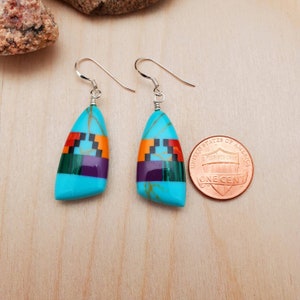 May include: A pair of turquoise and multi-colored inlay dangle earrings with silver ear wires. The earrings are about the size of a US penny.