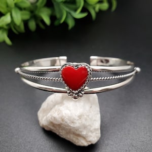 Op de afbeelding: Een zilveren armband met een rode hartbedel. De armband heeft een gedraaid touwontwerp en is aan &eacute;&eacute;n kant open.