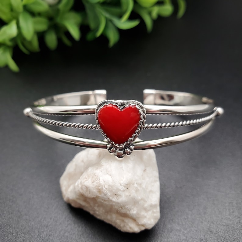 Op de afbeelding: Een zilveren armband met een rode hartbedel. De armband heeft een gedraaid touwontwerp en is aan &eacute;&eacute;n kant open.