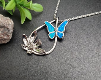 May include: A silver-toned pendant necklace featuring a butterfly with blue opal wings and a small clear gem. The pendant also includes a flower and is suspended from a delicate chain. The necklace is displayed on a black surface.
