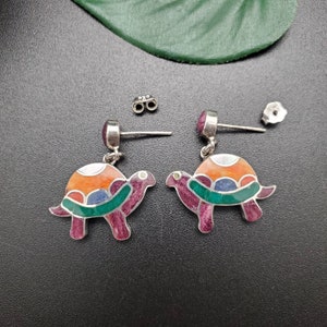 May include: A pair of silver turtle earrings with colourful enamel details. The turtles are a vibrant mix of orange, pink, green and blue.