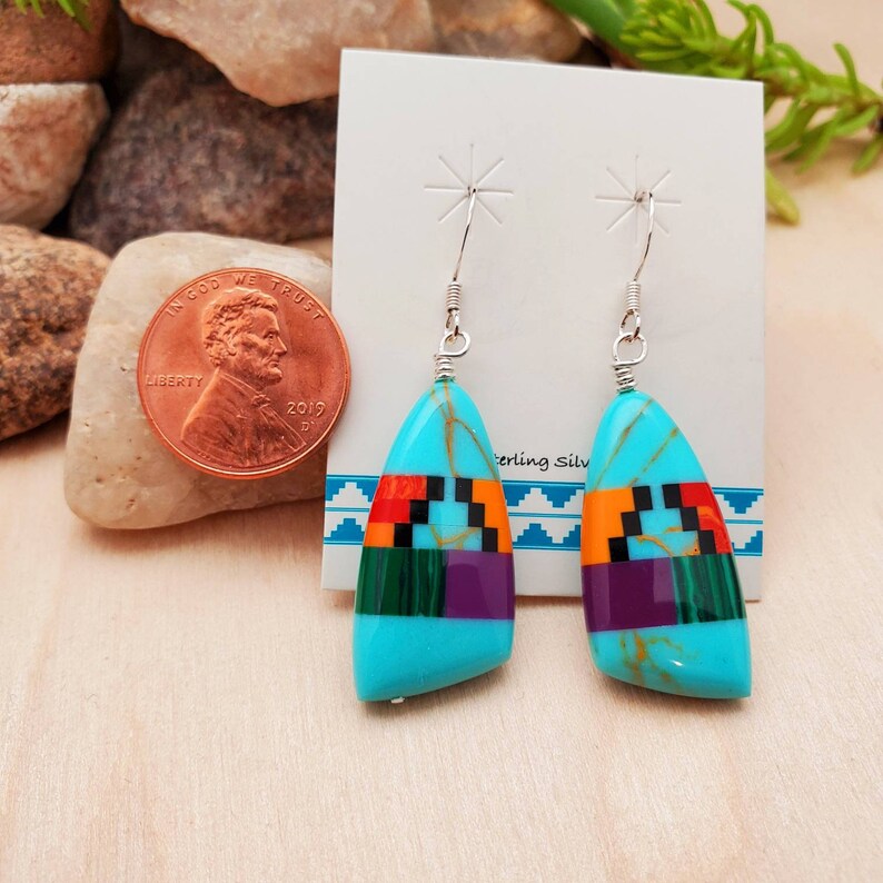 May include: A pair of turquoise dangle earrings with a geometric pattern of red, orange, green, purple, and black stones. The earrings are made with sterling silver and have a teardrop shape.
