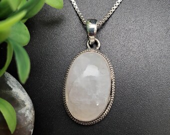 Puede incluir: Un collar de plata con un colgante ovalado de piedra preciosa clara. El colgante está engastado en un marco de plata con un borde en forma de cuerda y cuelga de una fina cadena de plata. El fondo es oscuro, con un toque de follaje verde.