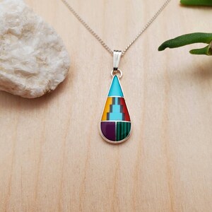 Puede incluir: Un collar con un colgante en forma de l&aacute;grima con incrustaciones de turquesa, amarillo, rojo, morado y verde. El colgante est&aacute; hecho de plata de ley y cuelga de una cadena de plata.