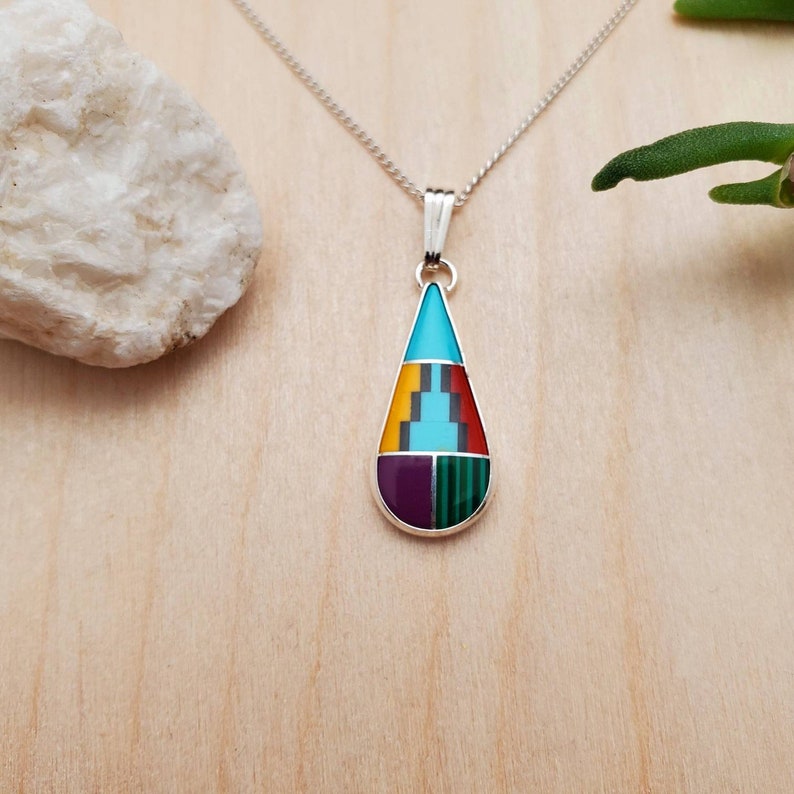 Puede incluir: Un collar con un colgante en forma de l&aacute;grima con incrustaciones de turquesa, amarillo, rojo, morado y verde. El colgante est&aacute; hecho de plata de ley y cuelga de una cadena de plata.