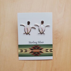May include: A pair of sterling silver earrings featuring a turtle design with a white opal inlay. The earrings are displayed on a white card with the text "Sterling Silver" and a Southwestern-style design.