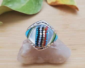 Op de afbeelding: Een zilveren ring met een vierkante steen in het midden. De steen is gemaakt van verschillende kleurrijke materialen, waaronder turquoise, rood en zwart.