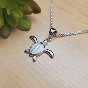May include: A silver chain necklace with a silver turtle pendant featuring an iridescent opal inlay. The pendant is on a wooden surface.