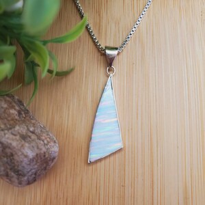 May include: A silver necklace with a triangular pendant. The pendant has a white, iridescent opal-like stone set in a silver frame, suspended from a fine chain. The background is a light brown wooden surface.