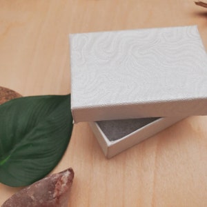 以下が含まれることがあります： 蓋にテクスチャパターンが施された白いギフトボックス。ボックスが開いており、内側は白い紙で裏打ちされています。