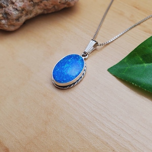 May include: A silver chain necklace with a blue oval opal pendant. The pendant is set in a silver bezel with a twisted rope design.