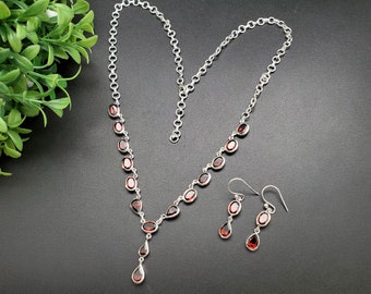 May include: A silver necklace and earring set with oval and teardrop-shaped red gemstones. The necklace has a delicate chain with oval stones and a teardrop pendant. The matching earrings have a similar design, with a hook closure. The set is displayed on a black surface.