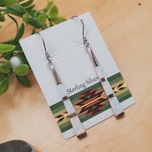 May include: A pair of dangle earrings with white opal stones set in sterling silver. The earrings are displayed on a card with a Southwestern-inspired design and the text "Sterling Silver".