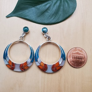 May include: A pair of silver hoop earrings with turquoise and orange inlay. The earrings are about the size of a US penny.