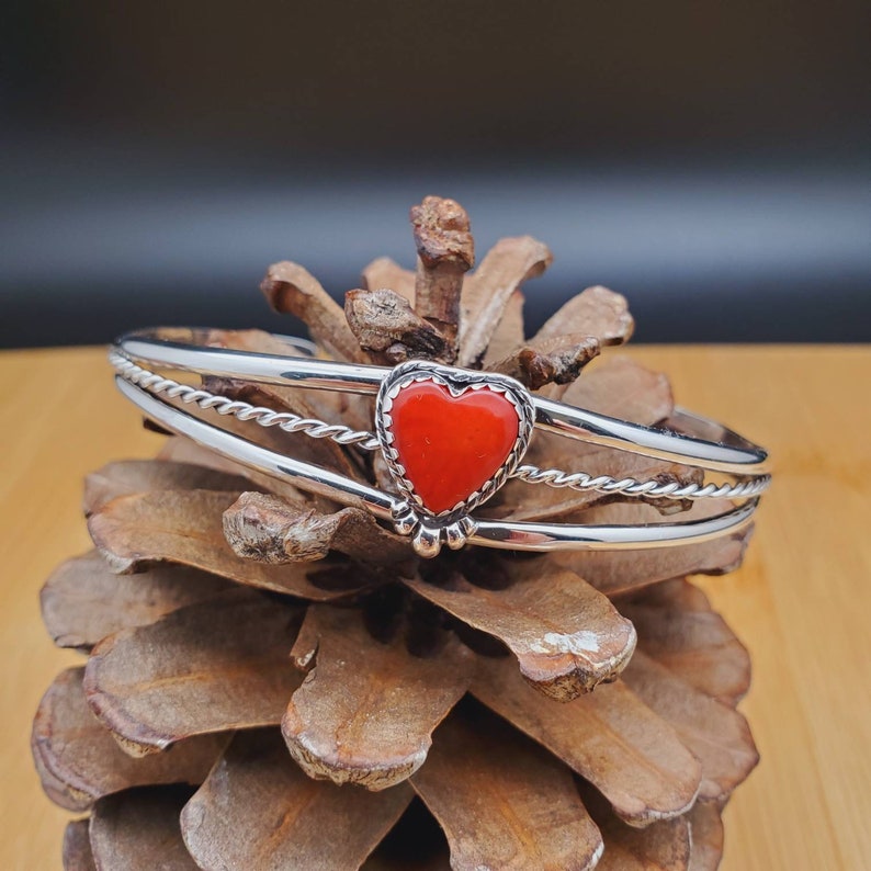 Op de afbeelding: Een zilveren armband met een rode hartvormige steen. De armband heeft een gedraaid draadontwerp.