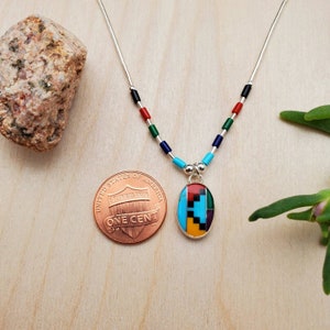 Puede incluir: Un collar de cadena de plata con un colgante que presenta un dise&ntilde;o geom&eacute;trico hecho de piedras turquesa, roja, negra, amarilla y verde. El colgante es aproximadamente del tama&ntilde;o de una moneda de un centavo de euro.