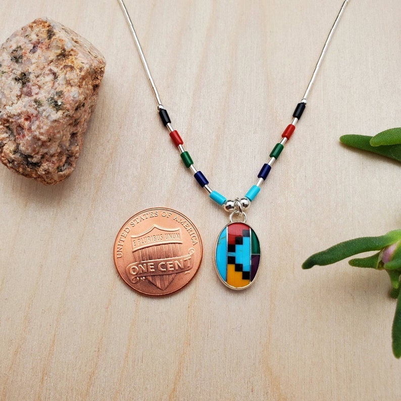 Puede incluir: Un collar de cadena de plata con un colgante que presenta un dise&ntilde;o geom&eacute;trico hecho de piedras turquesa, roja, negra, amarilla y verde. El colgante es aproximadamente del tama&ntilde;o de una moneda de un centavo de euro.