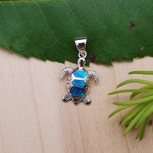 Può includere: Un ciondolo a forma di tartaruga in argento con una pietra preziosa di opale blu. Il ciondolo è su una superficie di legno con foglie verdi sullo sfondo.