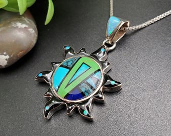 May include: A silver pendant necklace featuring a sunburst design. The pendant has a black enamel border with turquoise accents. The center of the pendant has a mosaic of turquoise, green, and blue stones. The necklace has a silver chain.